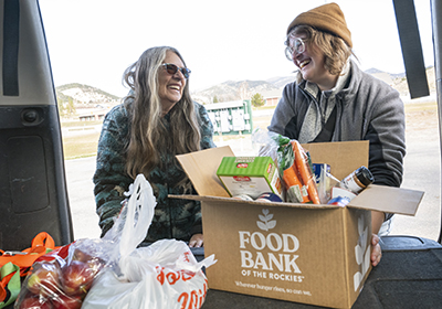 Two women smiling at one another while moving food out of a vehicle